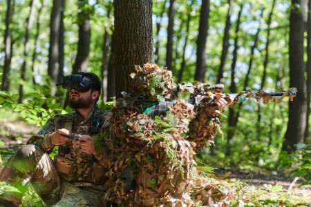 Foto de Un francotirador experto y un soldado que opera un dron con gafas VR diseñan estrategias y observan la acción militar mientras están ocultos en el bosque. - Imagen libre de derechos