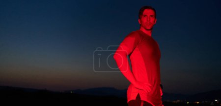 Foto de En la solemne oscuridad iluminada por un resplandor rojo, un atleta toma una postura segura, encarnando resiliencia y determinación después de completar un agotador maratón de un día de duración.. - Imagen libre de derechos