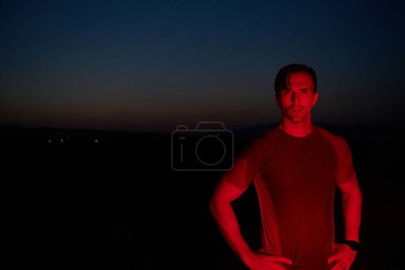 Téléchargez les photos : Dans l'obscurité solennelle éclairée par une lueur rouge, un athlète prend une pose confiante, incarnant résilience et détermination après avoir terminé un épuisant marathon d'une journée.. - en image libre de droit