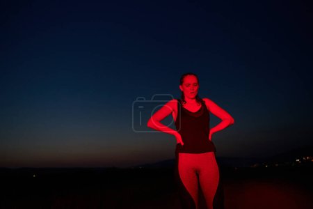 Téléchargez les photos : Après une course nocturne épuisante, un athlète prend une pose confiante, baignée dans la lueur rouge de l'environnement, montrant à la fois épuisement et détermination.. - en image libre de droit
