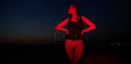 Photo for After a grueling nighttime run, an athlete strikes a confident pose, bathed in the red glow of the surroundings, showcasing both exhaustion and determination. - Royalty Free Image