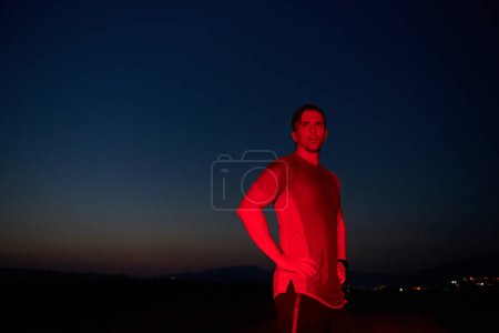 In der feierlichen Dunkelheit, die von einem roten Schein erhellt wird, nimmt ein Athlet eine selbstbewusste Pose ein und verkörpert Widerstandskraft und Entschlossenheit nach einem anstrengenden Marathontag..
