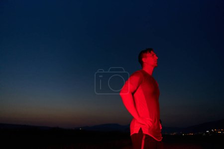 Dans l'obscurité solennelle éclairée par une lueur rouge, un athlète prend une pose confiante, incarnant résilience et détermination après avoir terminé un épuisant marathon d'une journée..