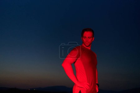 Photo for In the solemn darkness illuminated by a red glow, an athlete strikes a confident pose, embodying resilience and determination after completing a grueling day-long marathon. - Royalty Free Image