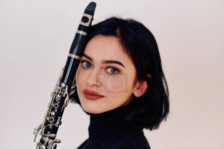 Téléchargez les photos : Une musicienne brune talentueuse met en valeur son art alors qu'elle tient gracieusement et joue de la clarinette sur un fond blanc immaculé - en image libre de droit
