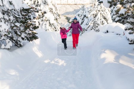Téléchargez les photos : Une mère et sa fille se précipitent le long d'un chemin enneigé serein, embrassant la beauté tranquille de leur escapade hivernale en montagne. - en image libre de droit