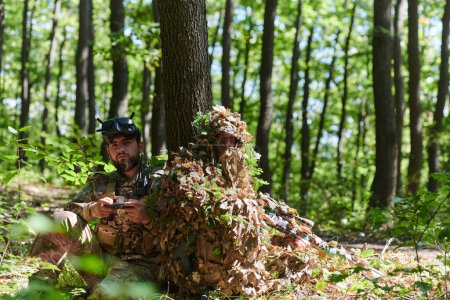 Foto de Un francotirador experto y un soldado que opera un dron con gafas VR diseñan estrategias y observan la acción militar mientras están ocultos en el bosque. - Imagen libre de derechos