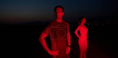 Téléchargez les photos : Dans l'obscurité solennelle éclairée par une lueur rouge, un athlète prend une pose confiante, incarnant résilience et détermination après avoir terminé un épuisant marathon d'une journée.. - en image libre de droit