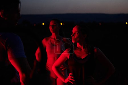 Foto de En las últimas horas de la noche, un diverso grupo de atletas exhaustos encuentran consuelo bajo un resplandor rojo, reflexionando sobre su viaje maratónico de un día y celebrando la camaradería en medio de la fatiga.. - Imagen libre de derechos