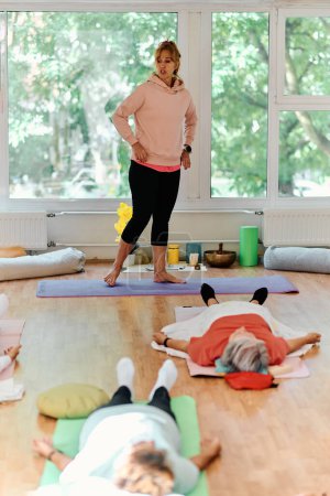 Téléchargez les photos : Un formateur qualifié supervise un groupe de femmes âgées pratiquant divers exercices de yoga, y compris le cou, le dos et les jambes, dans un espace ensoleillé, favorisant le bien-être et l'harmonie. - en image libre de droit