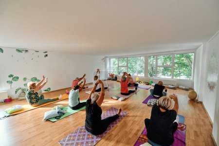 Foto de Un grupo de mujeres mayores realizan diversos ejercicios de yoga, incluyendo estiramientos de cuello, espalda y pierna, bajo la guía de un entrenador en un espacio iluminado por el sol, promoviendo el bienestar y la armonía.. - Imagen libre de derechos