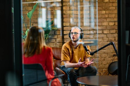 A gathering of young business professionals, some seated in a glass-walled office, engage in a lively conversation and record an online podcast, embodying modern collaboration and dynamic interaction
