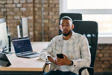  Afroamerikanischer Unternehmer macht eine Pause in einem modernen Büro, nutzt ein Smartphone, um in den sozialen Medien zu surfen, und fängt einen Moment digitaler Konnektivität und Entspannung inmitten seiner geschäftlichen Bemühungen ein