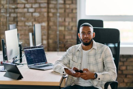 Téléchargez les photos : Un entrepreneur afro-américain fait une pause dans un bureau moderne, utilisant un smartphone pour naviguer sur les médias sociaux, capturant un moment de connectivité numérique et de détente au milieu de ses entreprises - en image libre de droit