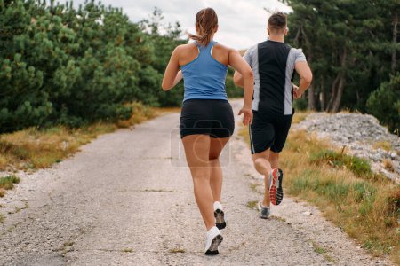 Téléchargez les photos : Un couple vêtu de vêtements de sport court le long d'une route pittoresque pendant une séance d'entraînement tôt le matin, profitant de l'air frais et maintenant un mode de vie sain. - en image libre de droit