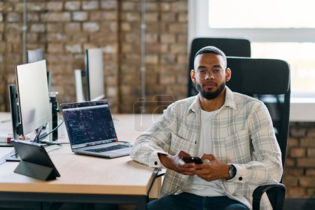  Afroamerikanischer Unternehmer macht eine Pause in einem modernen Büro, nutzt ein Smartphone, um in den sozialen Medien zu surfen, und fängt einen Moment digitaler Konnektivität und Entspannung inmitten seiner geschäftlichen Bemühungen ein