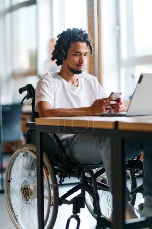 Ein afroamerikanischer Geschäftsmann im Rollstuhl macht eine Arbeitspause und benutzt sein Smartphone, während er in einem modernen Start-up-Coworking-Center sitzt, was Inklusion und Technologie widerspiegelt