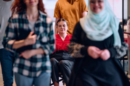 Téléchargez les photos : Les personnes qui parcourent un couloir dans le bureau vitré d'une start-up moderne, y compris une personne en fauteuil roulant et une femme portant un hijab. - en image libre de droit
