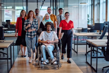 Photo for A diverse group of young business people congregates within a modern startups glass-enclosed office, featuring inclusivity with a person in a wheelchair, an African American young man, and a hijab - Royalty Free Image