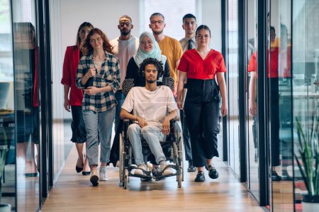 Foto de Un grupo diverso de jóvenes empresarios se congrega dentro de una moderna oficina cerrada en cristal de startups, con inclusión con una persona en silla de ruedas, un joven afroamericano y un hiyab. - Imagen libre de derechos