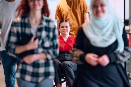 Foto de Personas que caminan por un pasillo en la oficina acristalada de una startup moderna, incluida una persona en silla de ruedas y una mujer con un hijab. - Imagen libre de derechos