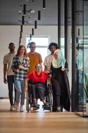  personas que caminan por un pasillo en la oficina acristalada de una startup moderna, incluida una persona en silla de ruedas y una mujer con un hijab.
