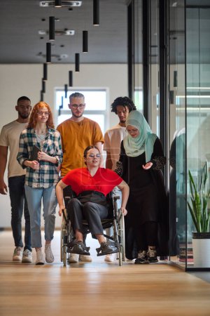 Foto de Personas que caminan por un pasillo en la oficina acristalada de una startup moderna, incluida una persona en silla de ruedas y una mujer con un hijab. - Imagen libre de derechos