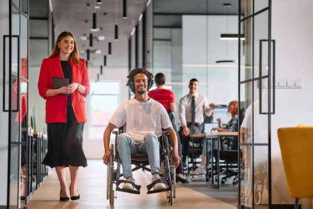 Une dirigeante d'entreprise avec son collègue, un homme d'affaires afro-américain handicapé, passe devant ses collègues qui travaillent dans des bureaux modernes.