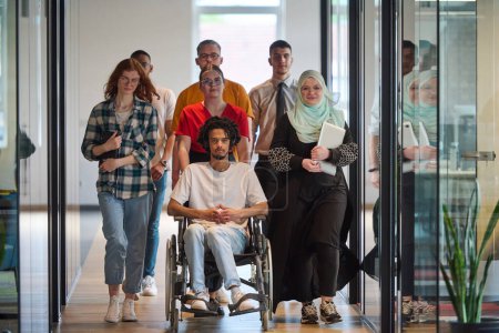 A diverse group of young business people congregates within a modern startups glass-enclosed office, featuring inclusivity with a person in a wheelchair, an African American young man, and a hijab