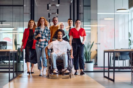Foto de Un grupo diverso de jóvenes empresarios que caminan por un pasillo en la oficina acristalada de una startup moderna, incluida una persona en silla de ruedas y una mujer con un hijab. - Imagen libre de derechos