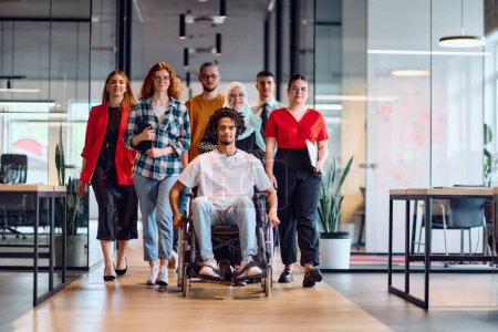 A diverse group of young business people walking a corridor in the glass-enclosed office of a modern startup, including a person in a wheelchair and a woman wearing a hijab.