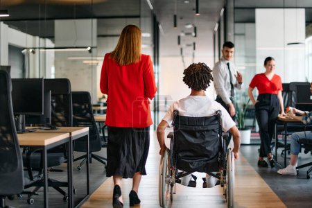 Una líder de negocios con su colega, un hombre de negocios afroamericano que es una persona discapacitada, pasa por sus colegas que trabajan en oficinas modernas.