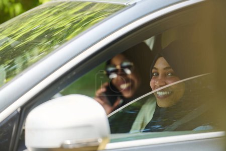 Foto de Dos mujeres musulmanas con hiyab conversan en un teléfono inteligente mientras viajan juntas en un coche a través de la. - Imagen libre de derechos
