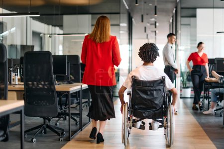Una líder de negocios con su colega, un hombre de negocios afroamericano que es una persona discapacitada, pasa por sus colegas que trabajan en oficinas modernas.