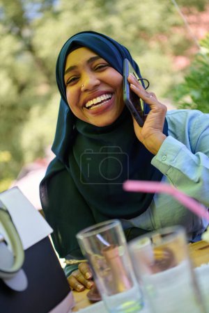 Téléchargez les photos : Une femme musulmane afro-américaine sereine en hijab engage une conversation paisible sur son téléphone portable au milieu de la beauté tranquille de la nature - en image libre de droit