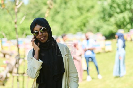  Muslimische Frau im Hijab aus dem Nahen Osten nutzt ein Smartphone, während sie ein kleines Imkereiunternehmen führt und dabei moderne Technologie mit traditionellen Praktiken verbindet