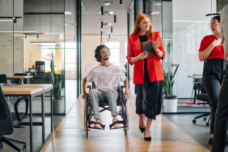 Téléchargez les photos : Une dirigeante d'entreprise avec son collègue, un homme d'affaires afro-américain handicapé, passe devant ses collègues qui travaillent dans des bureaux modernes. - en image libre de droit