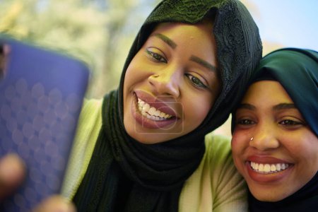 Téléchargez les photos : Deux femmes musulmanes du Moyen-Orient, parées de hijabs, capturent un moment d'amitié et de joie en prenant des selfies sur un smartphone alors qu'elles sont assises dans un cadre naturel. - en image libre de droit