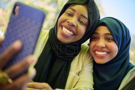 Téléchargez les photos : Deux femmes musulmanes du Moyen-Orient, parées de hijabs, capturent un moment d'amitié et de joie en prenant des selfies sur un smartphone alors qu'elles sont assises dans un cadre naturel. - en image libre de droit