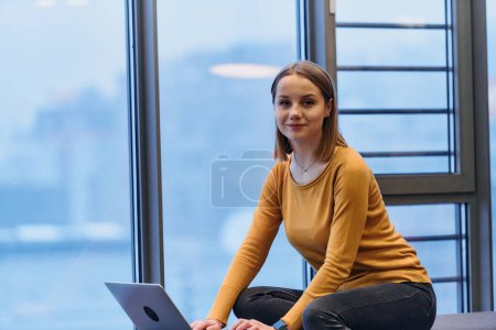 Téléchargez les photos : Une femme d'affaires utilise son ordinateur portable assis près de la fenêtre d'un grand bâtiment d'entreprise, offrant une vue pittoresque sur les toits de la ville comme toile de fond - en image libre de droit