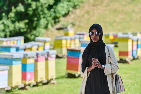 Mujer musulmana afroamericana en hiyab explora granjas de abejas naturales para la producción de miel, mezclando la tradición con la sostenibilidad en el campo sereno. 
