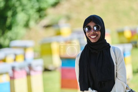 Afroamerikanisch-muslimische Frau im Hijab erkundet natürliche Bienenfarmen für die Honigproduktion und verbindet Tradition mit Nachhaltigkeit in der ruhigen Landschaft. 