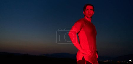 In der feierlichen Dunkelheit, die von einem roten Schein erhellt wird, nimmt ein Athlet eine selbstbewusste Pose ein und verkörpert Widerstandskraft und Entschlossenheit nach einem anstrengenden Marathontag..