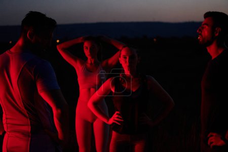 En las últimas horas de la noche, un diverso grupo de atletas exhaustos encuentran consuelo bajo un resplandor rojo, reflexionando sobre su viaje maratónico de un día y celebrando la camaradería en medio de la fatiga..