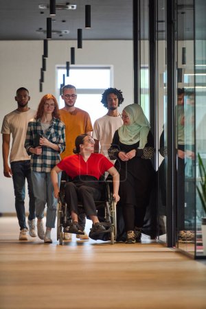  people walking a corridor in the glass-enclosed office of a modern startup, including a person in a wheelchair and a woman wearing a hijab.
