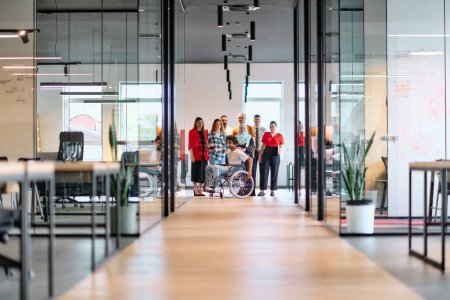 Un groupe diversifié de jeunes gens d'affaires marchant dans un couloir dans le bureau vitré d'une start-up moderne, y compris une personne en fauteuil roulant et une femme portant un hijab.