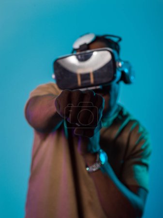En una escena vanguardista, un hombre afroamericano se involucra en juegos de realidad virtual de vanguardia, utilizando gafas VR para sumergirse en juegos de boxeo futuristas, contra un fondo azul vivo.