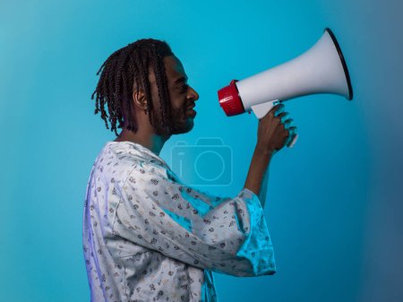Foto de El hombre afroamericano viste ropa tradicional, utilizando apasionadamente un megáfono contra un llamativo fondo azul, simbolizando su empoderamiento vocal y cultural en la búsqueda de la justicia social y - Imagen libre de derechos