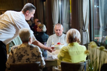 A courteous waiter provides impeccable service to an elderly family and their friends, creating a convivial atmosphere as they gather for a shared lunch, indulging in the diverse and delectable