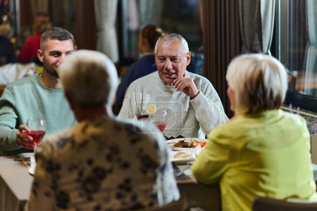 Eine Gruppe von Familienfreunden, bestehend aus einem jungen Enkel und älteren Menschen, teilen sich ein köstliches Abendessen in einem modernen Restaurant und veranschaulichen so das Konzept des gesunden Alterns durch generationenübergreifende